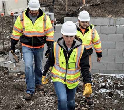 A Caucasian woman wearing a black hoodie, blue jeans, and safety vest is walking in front of a Caucasian man wearing brown pants and a safety jacket and a Caucasian man wearing a gray shirt on a work site.