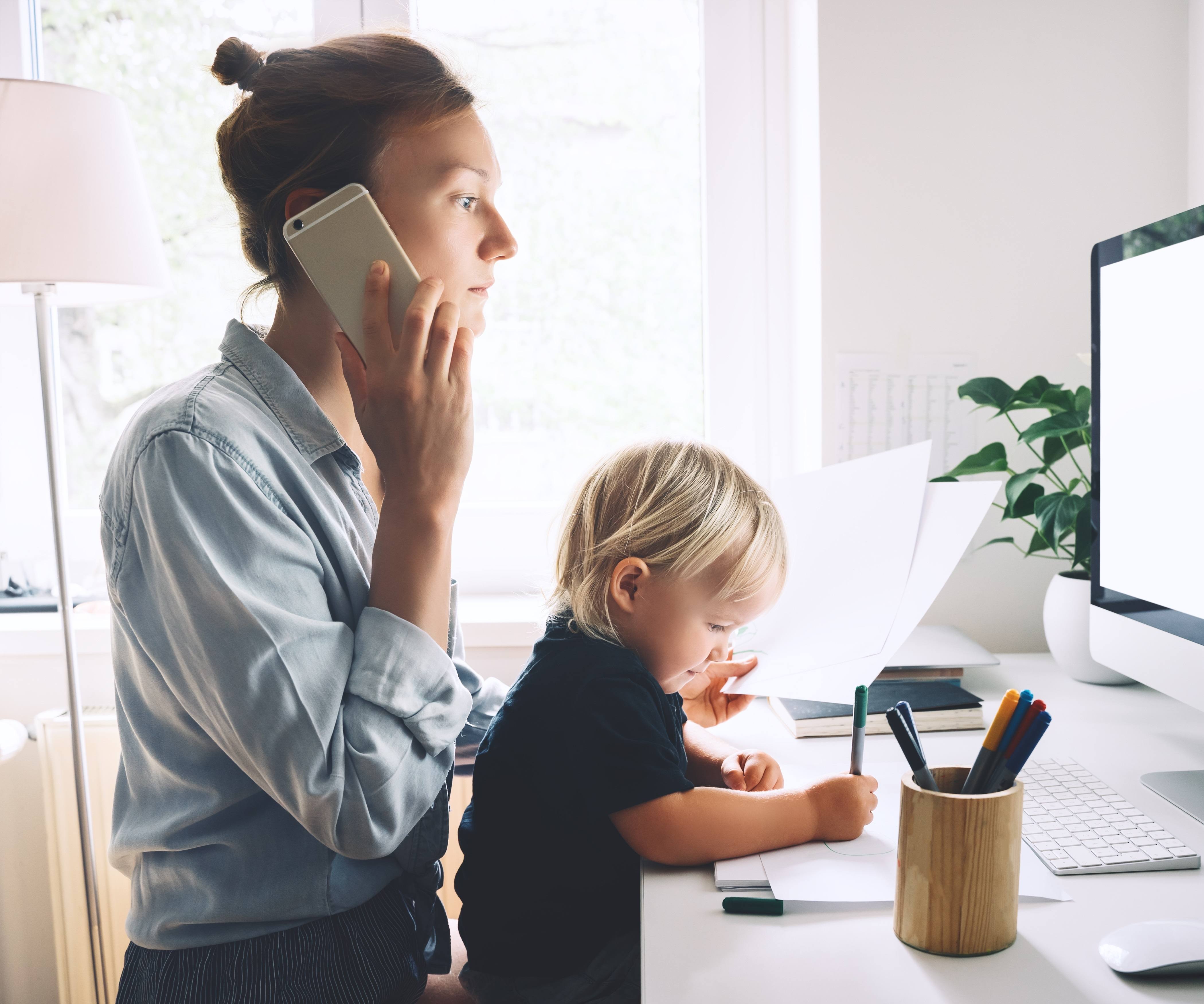 Modern mom balances between work and child home schooling on sick leave or quarantine. Mother works from home with kid. Woman responding on phone calls, working on computer with a toddler on her lap.